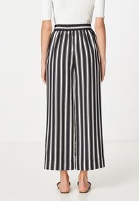 Žena v černobílých kalhotách s širokými nohavicemi a vertikálními pruhy s elastickým pasem, v kombinaci s otevřenými sandály na ploché podrážce.