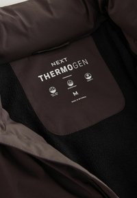 Eticheta pe o țesătură neagră și maronie închisă, arătând "NEXT THERMOGEN," mărimea M, cu iconițe pentru căptușeală termică, rezistență la apă și căldură, fabricat în Myanmar.