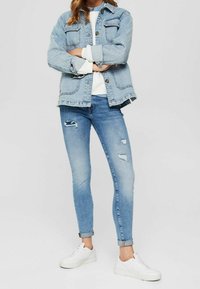 Chaqueta de mezclilla en azul claro con dos bolsillos en el pecho, llevada sobre una blusa blanca; combinada con unos vaqueros skinny en azul medio con desgastes y zapatillas blancas.