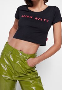 Camiseta negra corta con el texto "MISS SIXTY" en rosa, combinada con pantalones de tiro alto brillantes de color verde que presentan costuras y botones visibles.