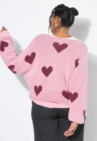Donna con capelli scuri raccolti in uno chignon, indossa un maglione rosa peloso con cuori bordeaux e pantaloni neri a righe sottili, di spalle con un braccio sollevato.