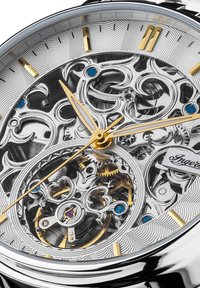 Montre squelette avec un boîtier en acier inoxydable argenté, un cadran transparent révélant des rouages complexes, des accents dorés et bleus, et un fond texturé.