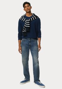 Marineblaues Langarmhemd mit einem gestreiften Strickpullover, der um den Hals gelegt ist, kombiniert mit hellblauen Jeans und blauen Sneakers.