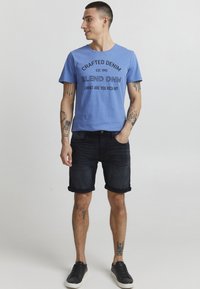 Blå bomulls T-skjorte med svart tekst, kombinert med mørke denimshorts med oppbrett; modell med svarte joggesko.