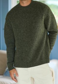 Olive green woolen sweater with a ribbed round neck, drop shoulders, and textured finish, paired with light-colored trousers.