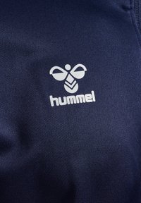 Weißes stilisiertes Bienenlogo und "hummel" Schriftzug auf dunkelblauem, strukturiertem Stoff, wahrscheinlich Sportbekleidungsstoff.