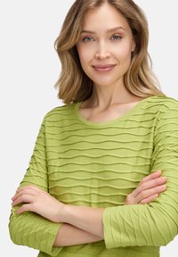 Hellgrüner Oberteil mit gewelltem Stoff, langen Ärmeln und rundem Ausschnitt. Weiches, leichtes Material mit bequemer Passform.