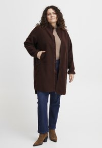 Abrigo de forro polar marrón con cremallera frontal, cuello alto y dos bolsillos frontales; combinado con jeans azules y botas de tacón marrones.