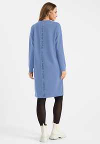 Vrouw in een blauwe jurk met lange mouwen met de tekst "FRIEDA NEW YORK" op de rug, gecombineerd met zwarte panty’s en witte enkellaarsjes.