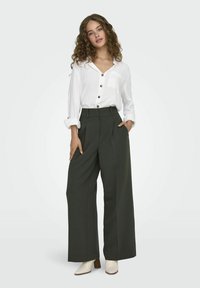 Hvit skjorte med knapper og lomme, kombinert med høytlivde, vide mørkegrønne bukser. Modellen har på lyse ankelstøvler.