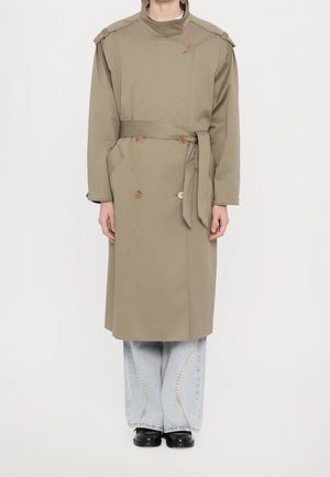 Person iført en lang beige trenchcoat med bælte over lyseblå vidde bukser og sorte sko mod en ensfarvet hvid baggrund.