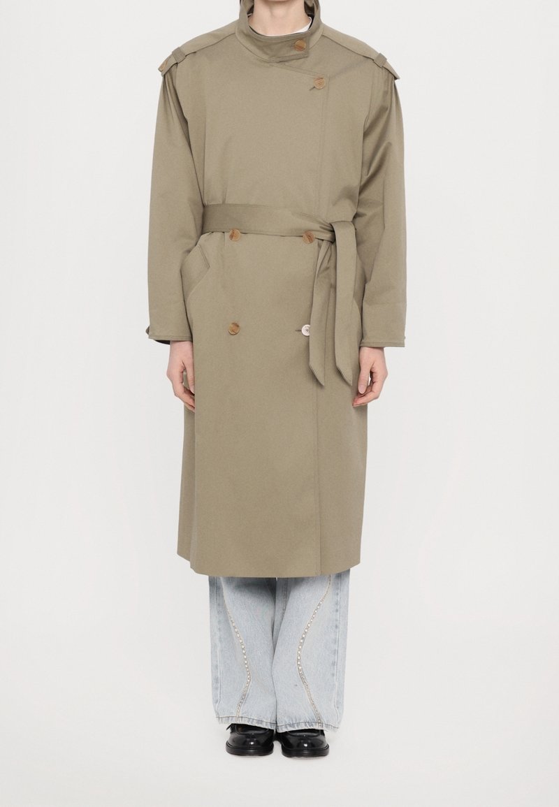 Personne portant un long trench-coat beige ceinturé sur un jean large bleu clair et des chaussures noires sur un fond blanc uni.