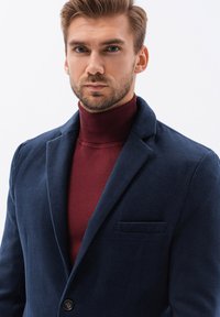 Mörkblå kavaj med mjuk textur, spetsig krage, en knapp och en framficka, i kombination med en burgundy polotröja.