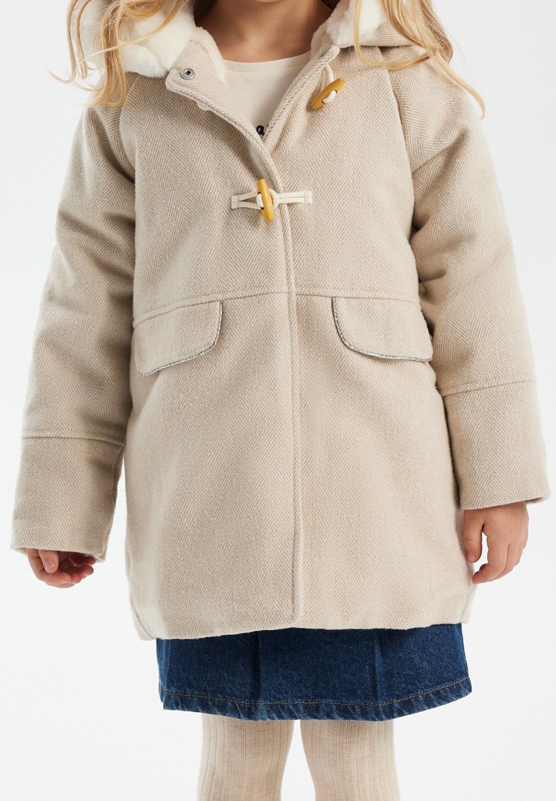 Enfant portant un manteau en laine beige avec des boutons à bascule, une jupe en jean et des collants crème sur un fond blanc uni.