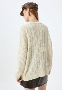 Beige Kabelstrickpullover mit geripptem Saum, locker sitzend und mit detaillierter Textur. Getragen mit einer braunen Mütze und einem Lederrock.