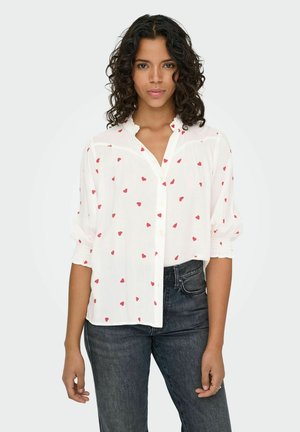 Chemise blanche à boutons avec un petit motif de cœurs rouges, col volanté et manches élastiques, associée à un jean en denim foncé.
