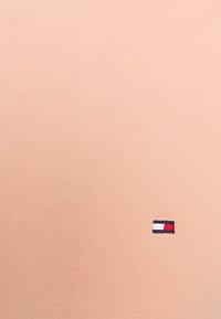 Jasno brzoskwiniowa, krótka koszulka z miękkiej bawełny o prążkowanej fakturze, z małą naszywką logo w kolorach czerwonym, białym i niebieskim na piersi.