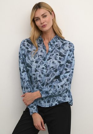 Blaues Blumen-Hemd mit Knöpfen, das ein aquarelliertes Muster zeigt, mit langen Ärmeln und einem Kragen, aus leichtem Stoff gefertigt. Schwarze Hosen werden getragen.