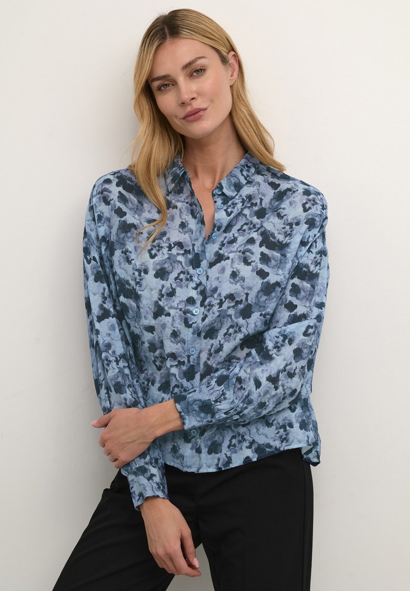 Camicia blu a fiori con bottoni, caratterizzata da un motivo acquerellato, maniche lunghe e colletto, realizzata in tessuto leggero. Sono indossati pantaloni neri.