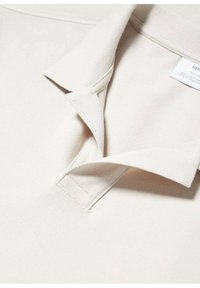 Polo beige avec un tissu texturé, doté d'un col classique, d'une patte de boutonnage pliée et d'une étiquette d'entretien visible sur le côté.
