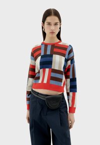 Pull court multicolore en rouge, bleu et marron avec un motif géométrique. Associé à un pantalon sombre et un sac ceinture noir.