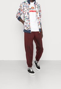 Färgglad, mönstrad zip-up-jacka med lappar, vit t-shirt med en logotyp, vinröda joggerbyxor och svarta sneakers med vita sulor.