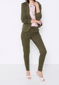 Olijfgroene blazer over een roze bloemen-top; bijpassende olijfgroene joggers; zachte stof; minimalistisch ontwerp; gecombineerd met lichtroze hoge hakken.