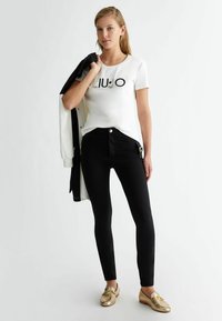 Balta īsām piedurknēm T-kreklu ar "LIUJO" logo, kas izšūts melnās un sudraba pērlītēs, apvieno ar melnām pieguļošām biksēm un zeltainiem sl�īdošiem apaviem.