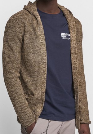Homme portant un cardigan en maille marron par-dessus un t-shirt bleu marine avec un texte blanc, ainsi qu'un pantalon beige avec un accessoire en chaîne sur la poche.