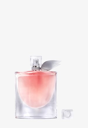 Flacon de parfum en verre carré avec liquide rose et ruban argenté noué près du spray, bouchon transparent retiré et posé à proximité.