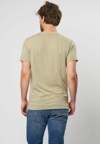 Beige pamut póló rövid ujjal és kerek nyakkivágással, párosítva kék farmernadrággal. Simább textúra, laza fazon, hátulról nézve.