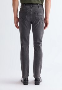 Man die strakke donkergrijze Wrangler-jeans en zwarte schoenen draagt, staande met zijn rug naar een effen lichte achtergrond gericht.