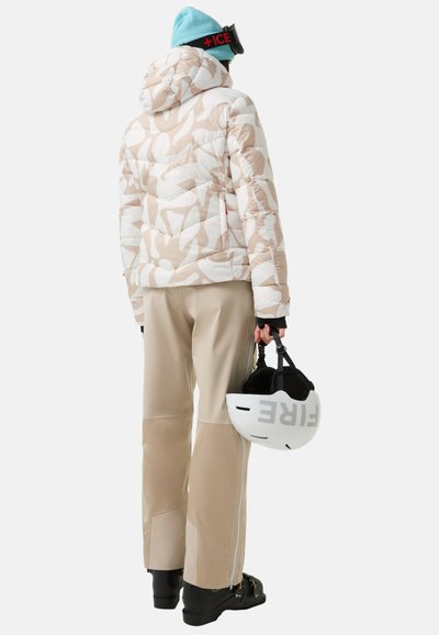 Polstret beige og hvid mønstret skijakke med hætte, kombineret med beige skibukser. Sorte støvler og en hvid hjelm holdt i hånden.