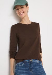 Braunes Langarmhemd mit Rundhalsausschnitt, kombiniert mit blauen Jeans. Das Model trägt eine grüne Mütze; glatte Textur, lässiges Design.