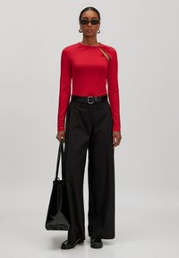 Roter Langarm-Top mit ausgeschnittenem Design, kombiniert mit schwarzen hochtaillierten weiten Hosen und einer glänzenden schwarzen Handtasche.