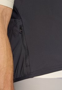 Haut de sport noir avec une poche zippée sur le côté, texture lisse et ourlet élastique. Le matériau est léger et extensible pour le confort.