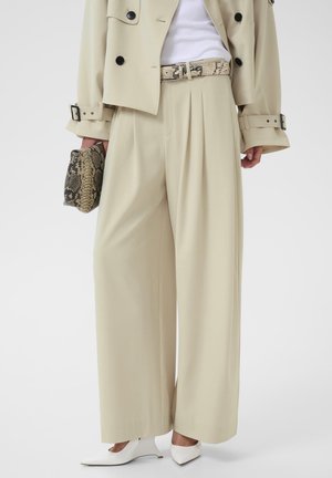 Naine, kes kannab beežikaid laiade säärega pükse, sobivat lühikest trentšmantlit, valgeid teravate otsadega kingi ja hoiab käes madu-nahkse mustriga käekotti.