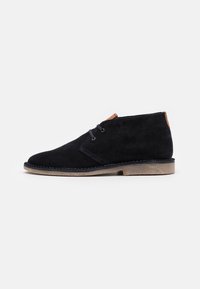 Stivale chukka in suede nero con punta rotonda, dettagli cuciti, accentuazione in pelle marrone chiaro al tallone e suola testurizzata marrone chiaro.