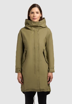 Vrouw draagt een knielange olijfgroene parka met capuchon en voorzakken, staand tegen een effen lichtgrijze achtergrond.