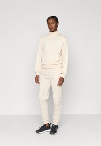 Beige träningsdräkt av mjukt tyg, med en jacka med dragkedja och hög krage samt matchande avsmalnande byxor. Svarta träningsskor med ränder.