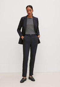 Blazer bleu marine avec revers crantés, haut rayé noir et blanc, pantalon bleu marine ajusté et mocassins noirs avec des accents métalliques.