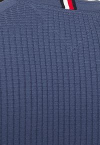 Blauer, gerippter Strickpullover mit hohem Kragen, der auf der Rückseite einen schmalen Streifenakzent in Rot und Weiß aufweist. Strukturierte Stoffdetails sichtbar.