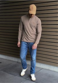 Camisa de manga larga beige, jeans azules y zapatillas blancas con detalles marrones. Gorra beige, de pie sobre una superficie de baldosas grises con una pared de tablones marrones.