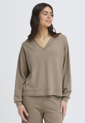 Kvinde med langt mørkt hår iført løstsiddende taupe V-hals sweatshirts og matchende bukser, der ser til venstre mod en ensfarvet baggrund.