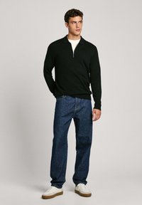 Schwarzer Reißverschluss-Pullover, lange Ärmel, weicher Stoff, kombiniert mit blauen Jeans im entspannten Schnitt und weißen Sneakers mit braunen Sohlen.