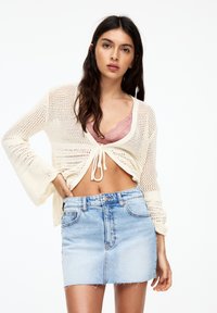 Knit creamfärgad cardigan med öppen väv, knytfrontdetalj, tillsammans med en ljusblå denimminikjol med fransat avslut. Rosa spets-bh under.