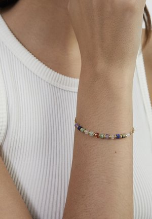 Nahaufnahme eines Handgelenks, das ein zartes Kettenarmband mit bunten runden Perlen trägt, vor einem Hintergrund eines weißen, gerippten ärmellosen Oberteils.