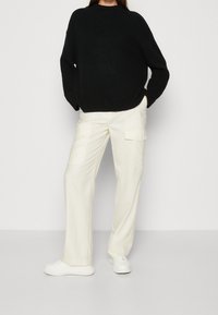 Schwarzer gerippter Strickpullover, oversized. Cremfarbene, weit geschnittene Hose mit Taschen, glatte Textur. Weiße Sneakers runden das Outfit ab.