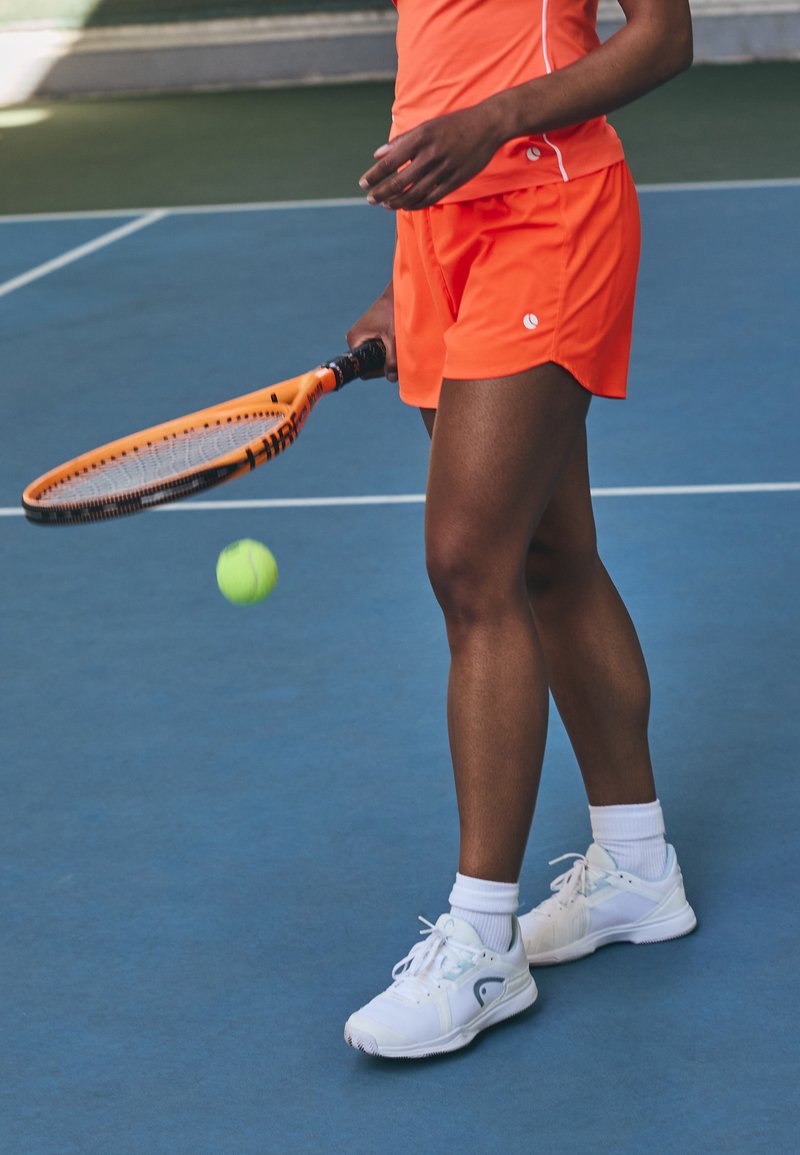 Tennisoutfit met een feloranje shirt en bijpassende shorts. De speler houdt een oranje racket vast en maakt zich klaar om een groene tennisbal te slaan.