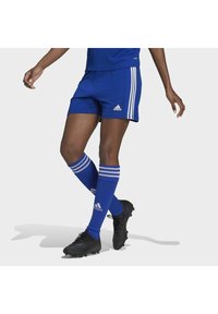 Osoba nosi plave Adidas nogometne šortse, odgovarajuće čarape do koljena i crne kopačke dok odlučno kroči naprijed na jednoličnoj pozadini.